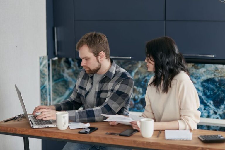 A couple reviews bills and finances at a desk with a laptop and papers. Indoor setting, focused on budgeting.